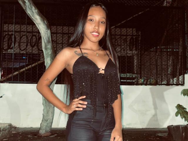 RiahannaJons with long black hair, wearing a black halter top with studs and black pants, standing with hand on hip, smiling, showing small tits and tattoo on left arm.