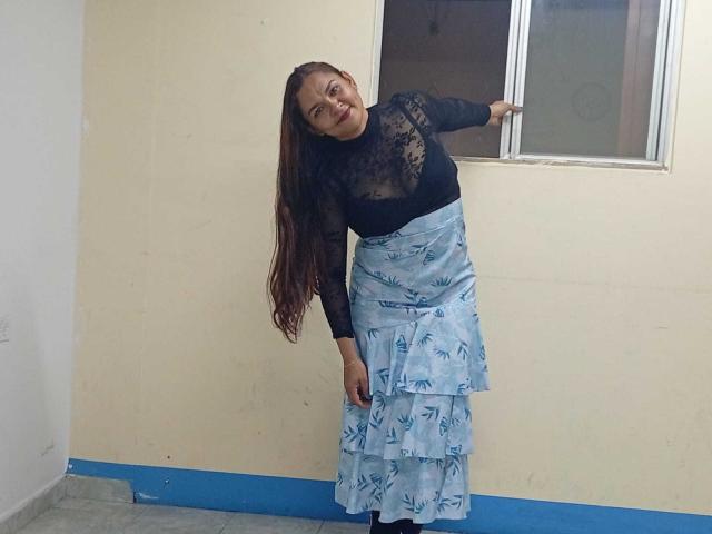 VivianBig with long brown hair, black lace top, and blue ruffled skirt, leaning against a window, smiling, wearing black shoes, standing on a blue-painted floor.
