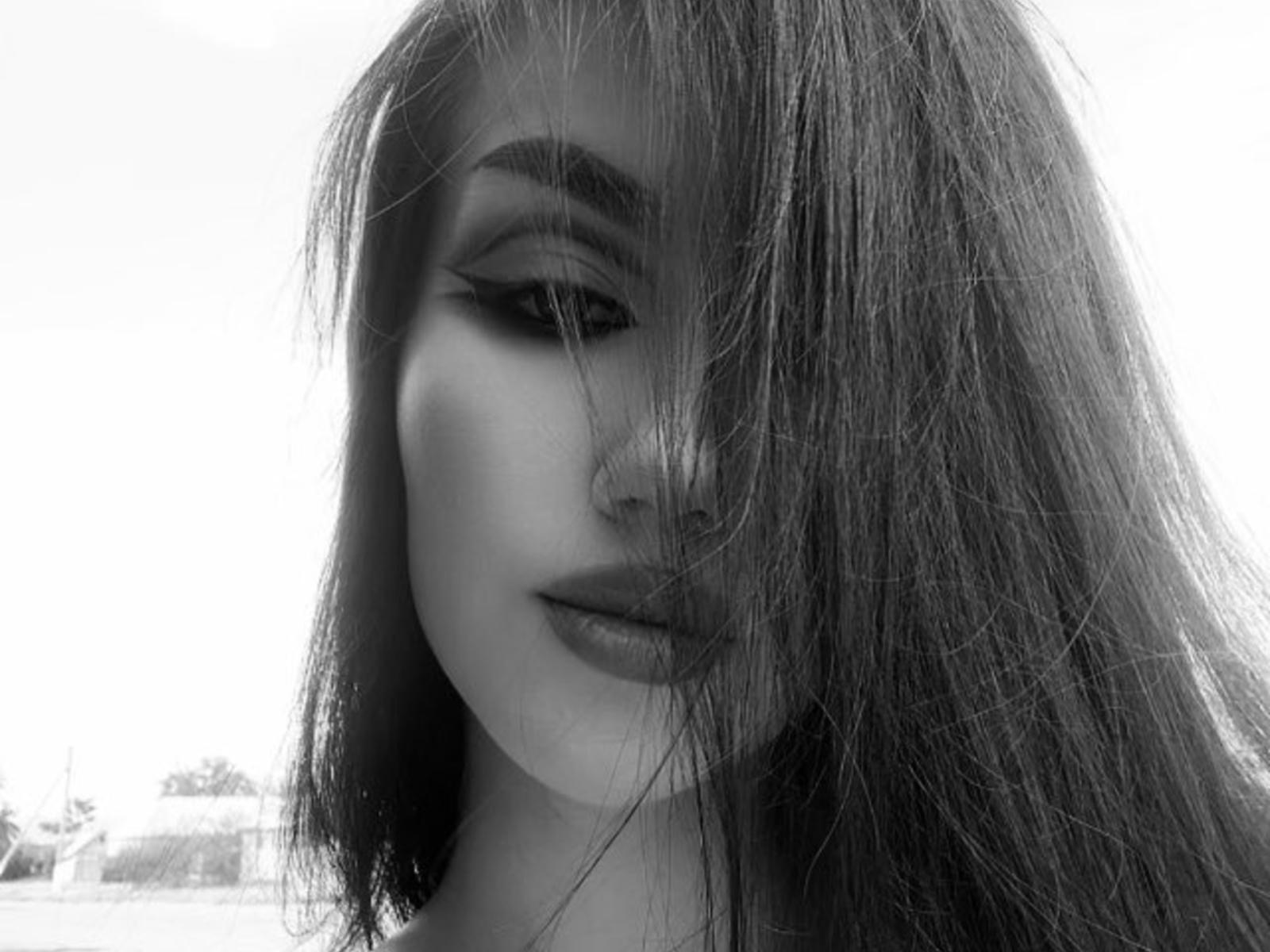Black-and-white photo of KarolMudge with long, messy hair covering one eye, thick eyebrows, and full lips, showing a slightly pouty expression. KarolMudge's face is centered, hair falls to side.