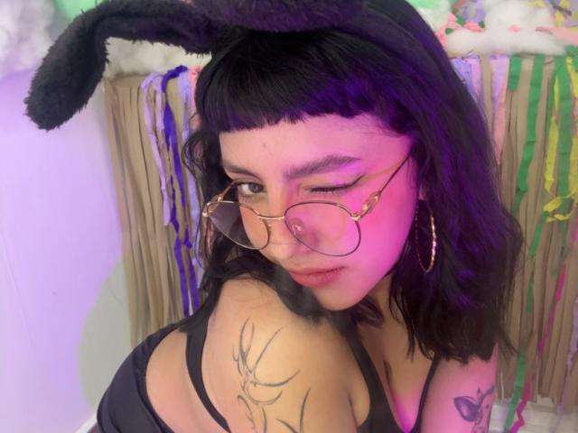 CuteH has dark, wavy hair with blunt bangs and wears black, fuzzy bunny ears. She's winking with one eye, wearing round, gold-rimmed glasses and large, gold hoop earrings. Her black tank top reveals a deer tattoo on her left shoulder. The purple and pink lighting casts a soft glow on her face. She has a small, round nose and full lips. Her skin is light brown. The background is blurred but includes colorful, hanging decorations. The image has a casual, playful vibe.