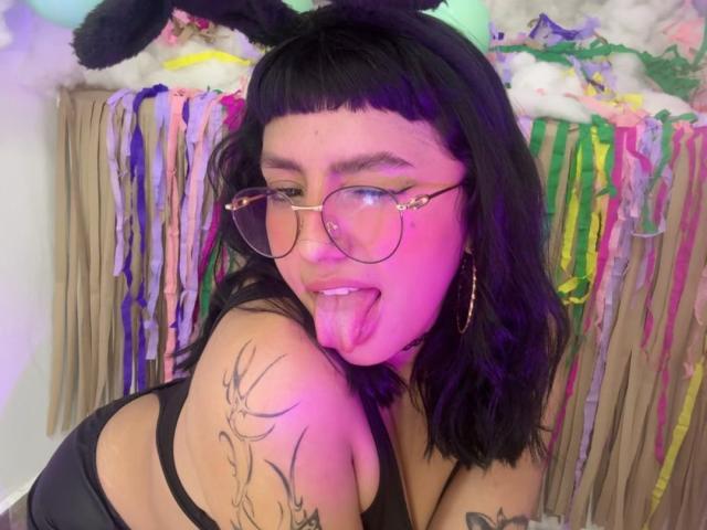 CuteH has black, wavy hair with bangs, wearing a black tank top, and has a tattoo of a deer on her left shoulder. She's sticking out her tongue, wearing gold-rimmed glasses with small pink studs, and has black bunny ears on her head. Her skin is light, and she's wearing large gold hoop earrings. The background is colorful with ribbons and fluffy decorations. The lighting is pink, highlighting her face and upper body.