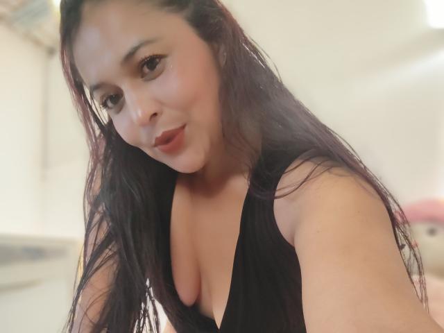 Close-up selfie of AfroditaLaDiosa with long, dark, slightly wavy hair, light brown skin, and subtle makeup. She has dark eyebrows, small red lips, and is looking slightly to the left. She wears a low-cut black top revealing moderate cleavage. The image is slightly blurry with a soft, warm light. Her expression is neutral with a slight, almost questioning, look. The background is out of focus, featuring light-colored surfaces and a hint of a pink object in the lower right.