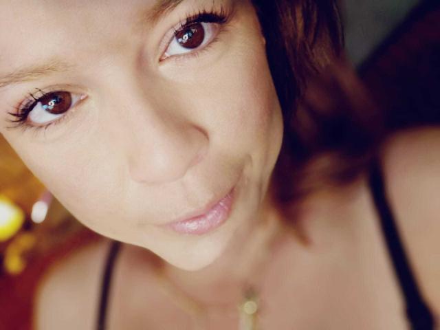 MoonXVelvet's face, showing brown eyes, long eyelashes, slight smile, and light brown wavy hair, wearing a black spaghetti strap top, with a gold necklace.
