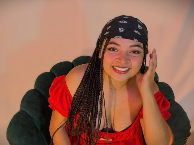ValerieDevereux with long braided black hair, red off-shoulder top, black bandana, smiling, hand touching ear, sitting on green chair, cute, medium tits, light skin, pink lips.