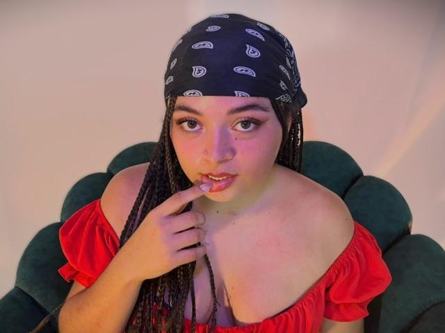 ValerieDevereux with long braids, wearing a black bandana, red off-shoulder top, and green chair, biting her finger, looking up with pink-tinted skin.