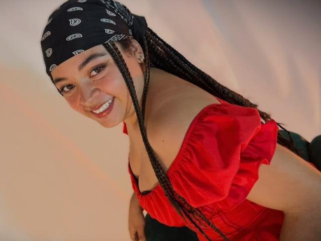 ValerieDevereux with long braids, smiling, wearing a red off-shoulder top and black bandana with white patterns, showing her ass slightly. She has a cute, attractive face.
