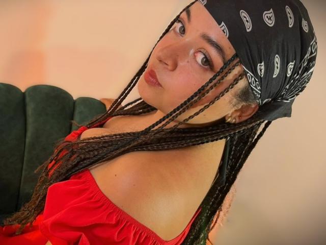 ValerieDevereux with long braided black hair, wearing a black bandana and red off-shoulder dress, looking over her shoulder with a pouty expression, showing her bare shoulders.