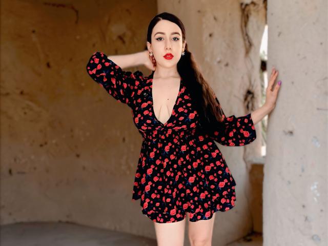 MabyLegrand stands in a dilapidated room with a rough, beige wall. She's wearing a black dress with pink cherry patterns, deep V-neckline, and puffy sleeves. Her right hand is in her hair, left hand on a pillar. She has long dark hair, red lipstick, and red earrings. Her skin is fair, and she has a curvy physique. The room has a broken, decayed feel with visible cracks. MabyLegrand's confident pose contrasts with the dilapidated background. She's centered in the frame.