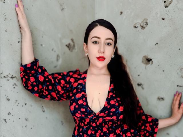MabyLegrand stands against a rough, gray concrete wall. She has fair skin, dark brown hair in a long ponytail, and wears bold red lipstick. Her dress is dark blue with a red floral pattern, deep V-neck revealing cleavage. Her right arm is raised, left arm is down. She has purple nails and small earrings. The composition is centered, with a direct gaze and slight smile. The texture of the concrete wall contrasts with her smooth skin and floral dress.