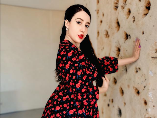 MabyLegrand stands against a rough, textured wall, facing the camera with a slight turn. She has long black hair, fair skin, and red lipstick. She wears a black dress with a red cherry pattern and touches the wall with her right hand. Her left hand rests on her hip. She has purple-painted nails and a small nose ring. Her expression is confident and slightly sultry. The image has a modern, slightly provocative style.