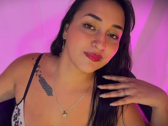 TatianaSokolova with glossy red lips, black hair, and a feather tattoo on her chest, wearing a white and black patterned top, silver necklace, and ring, with pink lighting.