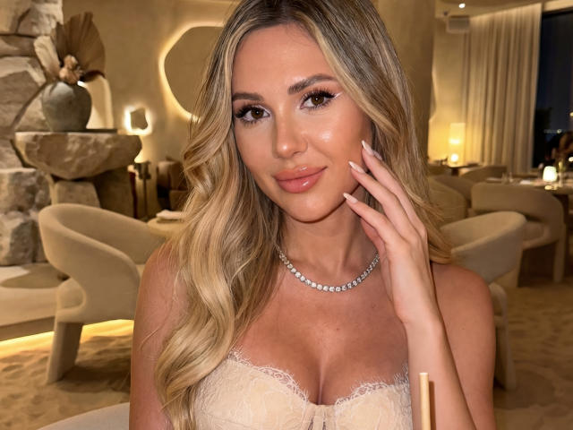 NensySafrons has long, wavy blonde hair and a light tan complexion. She's wearing a sheer, lace strapless top that reveals her cleavage. Her right hand is raised to her face, with manicured nails. She has a subtle smile and is wearing a silver necklace. The lighting is warm, highlighting her facial features. The background includes a stone structure and soft, beige decor. The image is well-composed, focusing on NensySafrons' face and upper body.