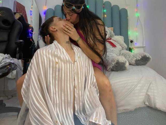 AnjaliRohan, long black hair, in pink lingerie, kisses a guy with short brown hair, in a striped shirt, sitting on a bed with a giant white stuffed animal.