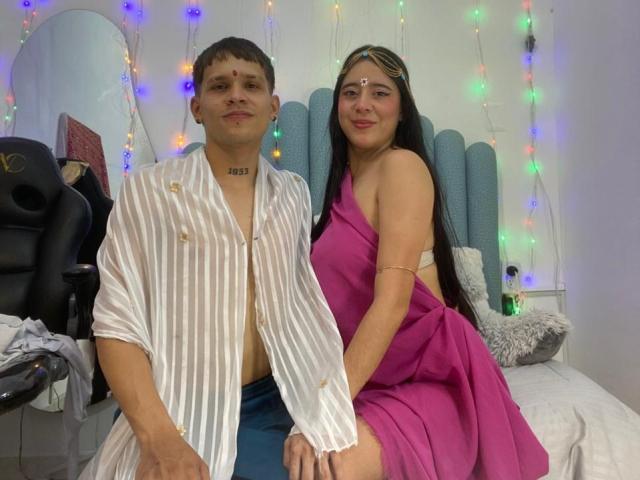 A young man with short brown hair and a tattoo on his neck sits beside AnjaliRohan, who has long black hair and a forehead dot. He wears a white striped shirt and blue pants, while she is in a pink dress with a gold arm cuff. Multicolored string lights are in the background. The man is on the left, AnjaliRohan on the right, both sitting on a bed.