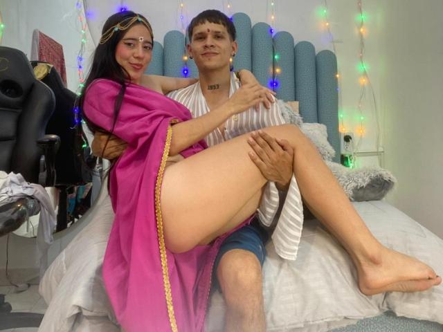 AnjaliRohan with long black hair and fair skin sits on a bed with a white-striped shirtless man. She wears a pink robe with yellow trim, exposing her legs. He has short brown hair, a tattoo on his neck, and wears blue shorts. She rests her arm on his shoulder, smiling. The bed has a blue padded headboard, and colorful string lights are in the background. The room has a black chair and scattered items. The composition is casual and intimate.