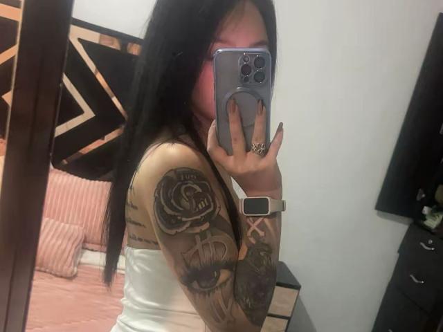 ViolettaSquirt with tattoos on arms, wearing white tank top, taking mirror selfie with iPhone, showing off tattoos, long black hair, pink face, ring on finger, wearing smartwatch.