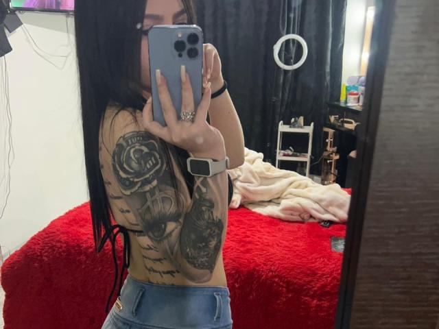 ViolettaSquirt, met lang zwart haar, onthuld, die grote tatoeages aan haar rechterkant laat zien, houdt een telefoon vast in een spiegelzelfportret, draagt blauwe jeans en een smartwatch..