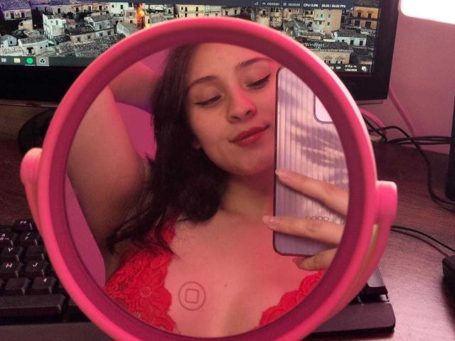 A selfie in a pink circular mirror shows AnnieLamberth with closed eyes, a slight smile, and long dark brown hair. She's wearing a red lace top, revealing a small tattoo of a circle with a line below it on her right chest. Her right arm is raised, and she holds a silver phone with a ridged back. A computer screen with a cityscape background is visible in the background.