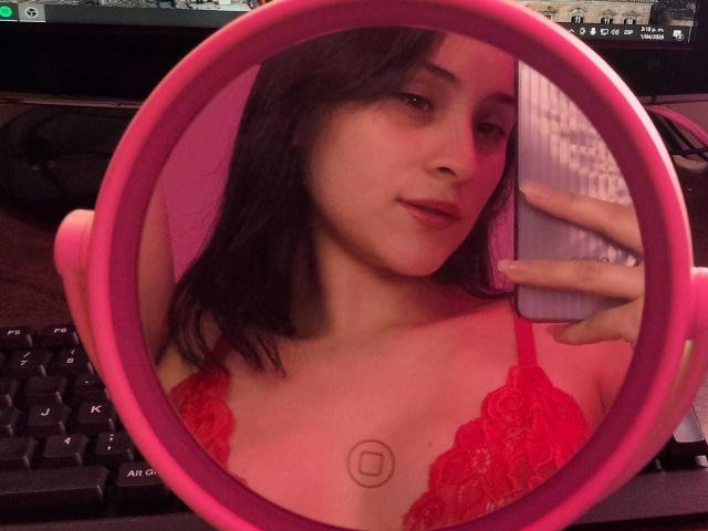 A close-up selfie in a pink circular mirror. AnnieLamberth, with fair skin and dark brown hair, looks into the mirror. She wears a red lace bra, showing a small, circular tattoo below her cleavage. Her right hand holds a white phone. The reflection is centered in the mirror, with her face and upper chest visible. The mirror's pink frame contrasts with her red bra and the black keyboard in the background. The image has a casual, intimate feel.