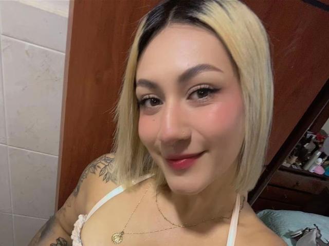 EmilyRobertts has blonde and black hair, tattoos on her arm, wearing a white lace top, pink lipstick, and a gold necklace, smiling with pink cheeks.