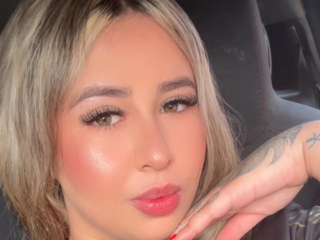 BelaSexyy's face, blonde hair, pink lipstick, subtle blush, tattoo on right hand, long eyelashes, brown eyes, red nail polish, slightly pouty lips, soft light, car interior.