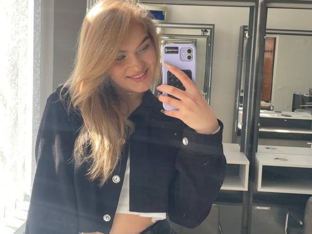 AlinaLemon with long blonde hair, smiling, holding a purple phone, wearing a black shirt, showing her tits, in a bright room, taking a mirror selfie.