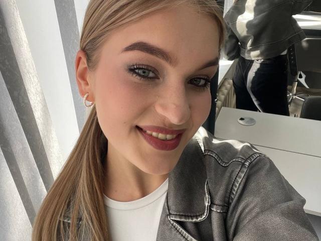 AlinaLemon has fair skin, blonde hair in a ponytail, blue eyes, dark red lipstick, white shirt, gray denim jacket, and a silver hoop earring, smiling at the camera.