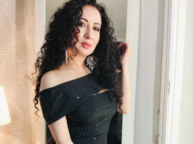 SamanthaAntolin stands in a room, facing slightly to the right. She has long, curly black hair and is wearing a black off-the-shoulder dress with small, glittery patterns. Her right hand touches her hair. She has light skin and wears large, floral-shaped earrings. The dress has a small, horizontal cutout on the left side. The light from a lamp on the left side casts a warm glow. The image is well-lit, emphasizing her facial features and dress texture.