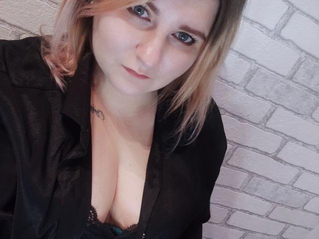 MinistraX with light brown hair, blue eyes, and slight pink lipstick, wearing a black shirt, exposing her tits in a black lace bra, against a white brick wall.