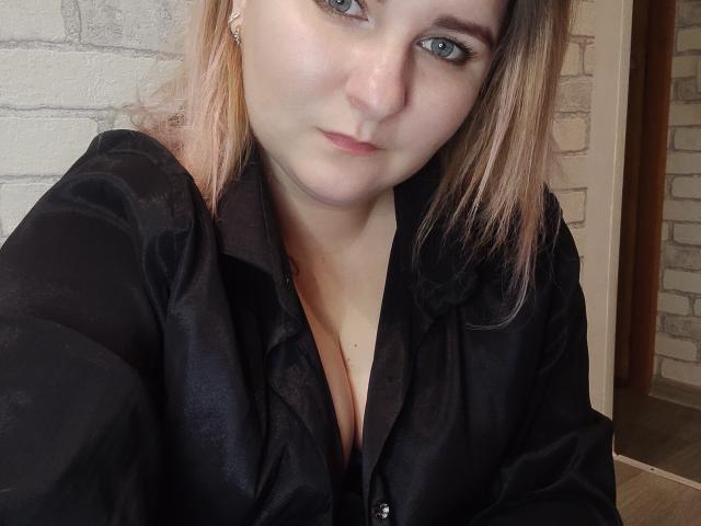 MinistraX with pale skin, blonde hair, and blue eyes, wearing a black, slightly open, satin shirt showing her cleavage, taking a selfie, looking directly at the camera.