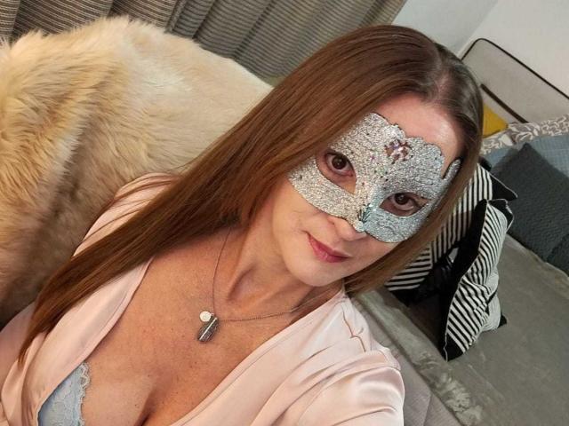 SabrinaMilf with long brown hair, wearing a sparkly silver masquerade mask, pink satin robe, and a blue lace bra, showing her tits, sitting on a bed.