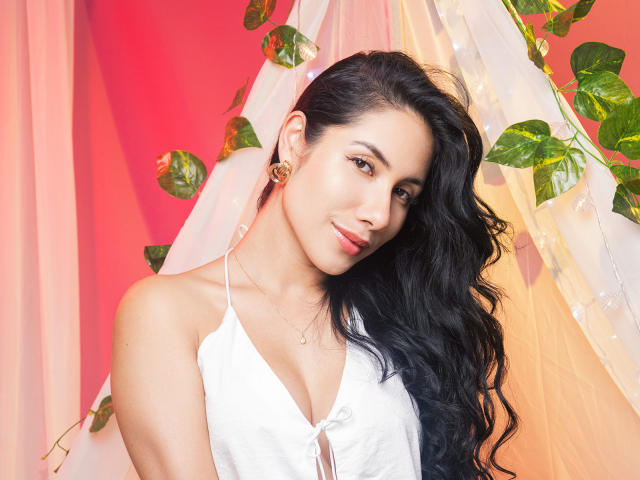 ArianaRogers with long black hair, white halter top, small gold earrings, and a subtle smile, showing her tits in a pink-lit setting with green leaves and white drapes.
