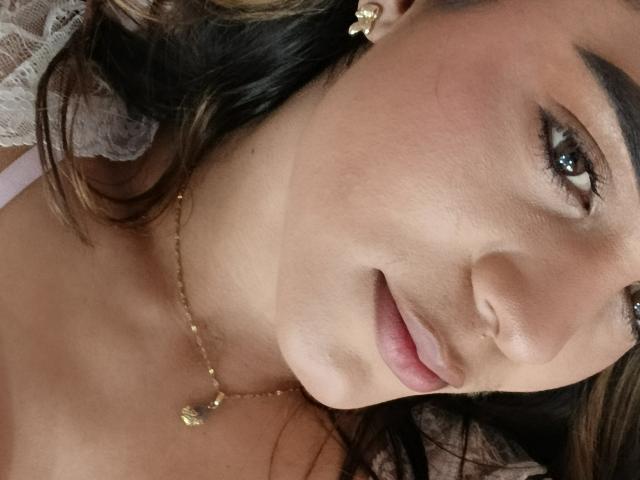 Close-up of AliceCoolenk's face. She has light brown skin, dark wavy hair with blonde highlights, and is looking slightly to the right. Her eyebrows are dark and well-defined, and she has light brown eyes with subtle eyeliner. She's wearing a small gold necklace with a leaf-shaped pendant and a gold cross earring in her right ear. Her lips are closed in a slight smile. The lace in the upper left corner is white and delicate. The composition focuses on her face, with a soft, natural light.