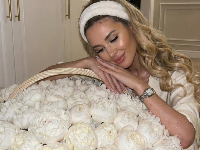 NensySafrons with long, wavy blonde hair, wearing a white headband and satin blouse, rests her face on a basket of white peonies. Her eyes are closed, and she has a slight smile. She's wearing a silver watch on her right wrist. The basket is filled with densely packed, fresh white peonies. The image has a soft, warm light. Her skin is slightly shiny.