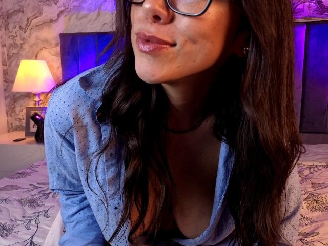Mia Reed, avec ses cheveux bruns ondulés, ses lunettes et ses lèvres brillantes, porte une chemise bleue à pois et dévoile légèrement sa poitrine dans un haut à décolleté bas. Chambre avec un éclairage violet et jaune..