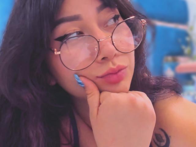 Genesiss, a young woman with long, wavy black hair, wears round, gold-rimmed glasses and has blue lipstick. She rests her chin on her hand, showcasing blue nail polish. Her tattoo, a black circular design on her right shoulder, is partially visible. The soft focus and warm, pinkish light highlight her relaxed, confident expression. The image has a slightly blurred, blue background.
