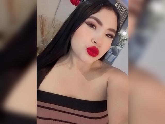 OliviGrace with long black hair, red lipstick, and a strapless, striped, brown dress, puckering her lips. She has bold eyebrows and a slight smile. Her tits are not visible.