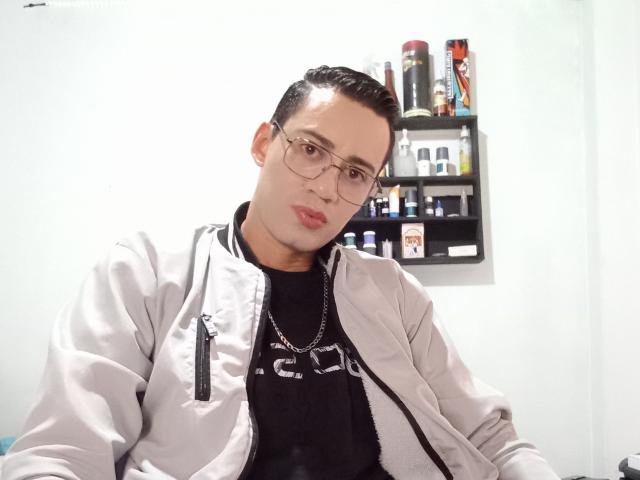 LucianoZ, nerdy-looking dude with glasses, light skin, short black hair, wearing a gray jacket, black shirt, and chain, sitting, slightly leaning forward, looking at camera.