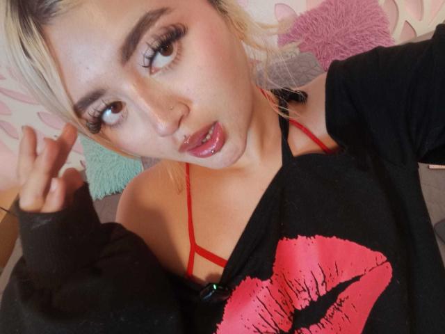 Close-up photo of LyaMoon with blonde hair, light skin, and brown eyes. She has thick, arched eyebrows, long, dark eyelashes, and a small nose ring. She wears a black sweatshirt with a red kiss print and a red strap visible from a thin top. Her mouth is slightly open with a glossy, pink lip. She holds up her right hand, showing a peace sign. The background has pink and green patterned pillows. The composition focuses on her face and upper body. The texture of the sweatshirt is soft and slightly worn.