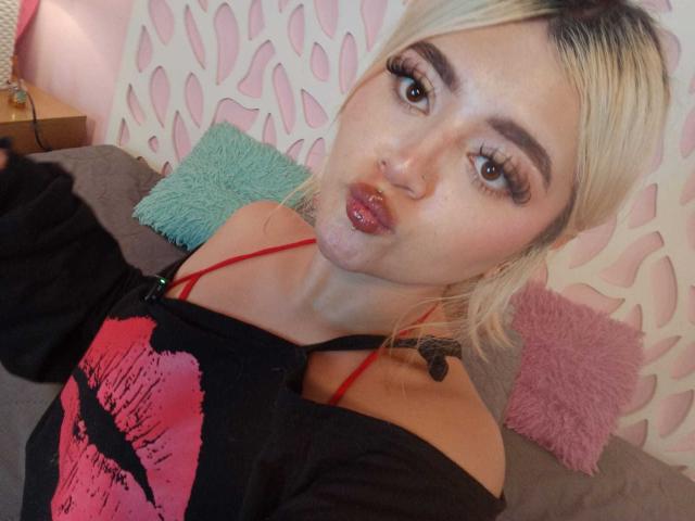 LyaMoon with blonde hair, pouty lips, and long lashes, wearing a black off-shoulder top with red straps, showing a pink lip print, and a small nose ring.