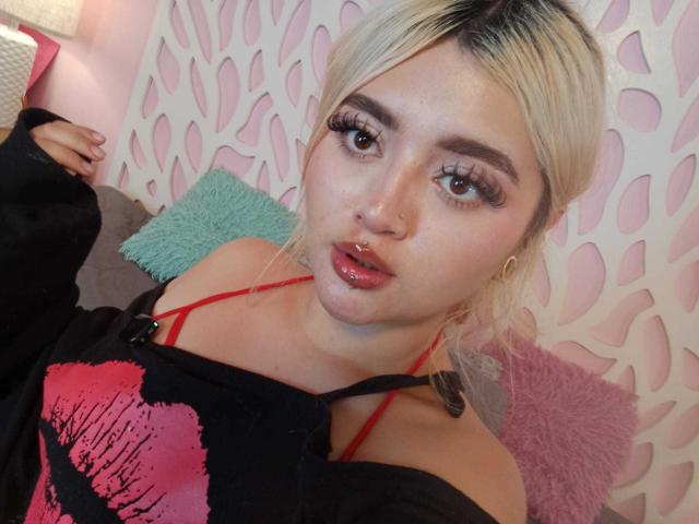 LyaMoon has platinum blonde hair, parted in the middle, and wears dramatic long eyelashes. She has a small nose ring in her right nostril and slightly parted glossy pink lips. She wears a black off-shoulder top with a red strap and a pink graphic. Her fair skin is smooth, and her eyebrows are dark and well-defined. She's seated on a gray couch with a green and pink pillow. The background has a white, leaf-patterned wall. The image is a close-up selfie.