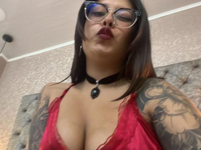Close-up of GabyJoy with medium-length dark brown hair, wearing glasses with thick black frames, and deep red lipstick. She is pouting her lips and looking down slightly. GabyJoy is dressed in a low-cut red lace top that reveals her large breasts and a black choker with a small black pendant. Her tattooed right arm is partially visible, showing intricate black and gray designs. The texture of the lace is delicate and slightly translucent. The image is slightly blurred, adding a soft, almost hazy effect to the overall composition. The lighting is even, highlighting her tattoos and the lace's texture.