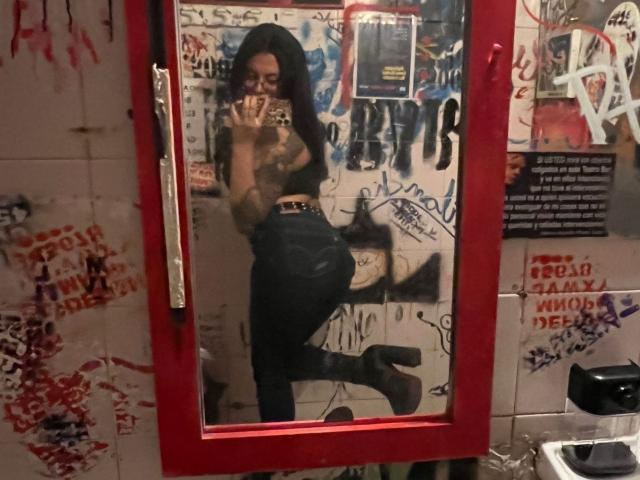 GabyJoy with long black hair, glasses, and a tattooed chest, taking a mirror selfie in a grungy room, wearing tight jeans and high-heel boots, showing her ass.