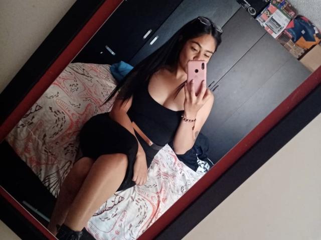 StellaBeckker in a black tube top and skirt, showing her tits and ass in a mirror selfie, holding a pink phone, with long black hair and a bracelet.
