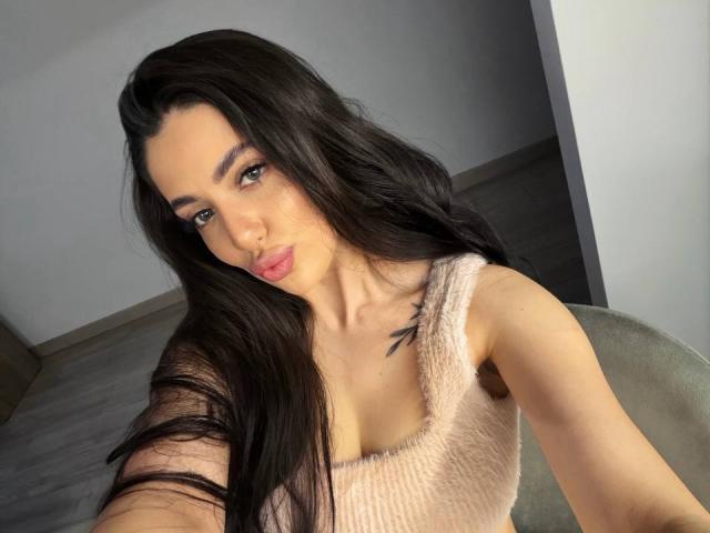 ArielGemme with long black hair, pink fuzzy top, pouty pink lips, green eyes, small tits, tattoo on right shoulder, taking selfie, gray chair, wooden floor, white wall.