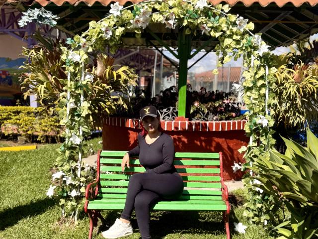 KatherineToro sitting on a green wooden bench with red arms, wearing black clothes and a white cap, under a floral arch in a sunny garden.