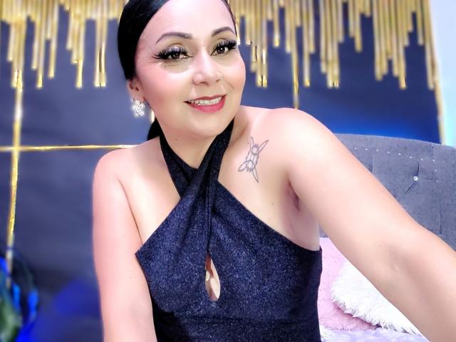 Zaria69 with black hair, wearing a sparkly black halter dress, showing her tits with a keyhole cutout, smiling, tattoo on left shoulder, gold earrings, and a pink pillow.