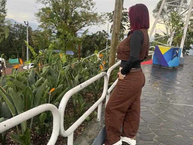 KatherineToro with red hair, brown pantsuit, black long-sleeve top, standing by white railing, looking away, large ass, holding white bag, near bird of paradise flowers, outside.