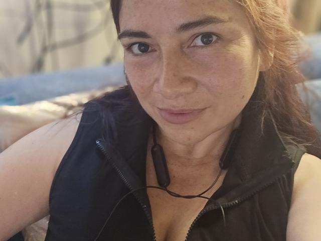 AlessiaRed, a brunette woman with freckles, wearing a black zip-up vest, looking directly at the camera with a slight smile, black earbuds around her neck.