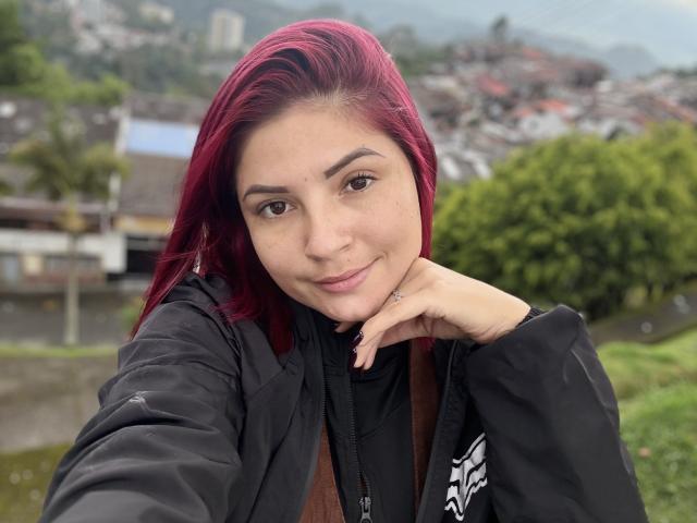 KatherineToro with red hair, black jacket, and brown shirt, smiling with chin on hand, brown eyes, light skin, in a green landscape, looking at camera, casual selfie.